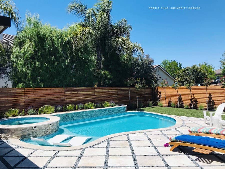 Sunny backyard with a pool, hot tub, and striped lounge chair, surrounded by trees and a wooden fence on a bright, clear day