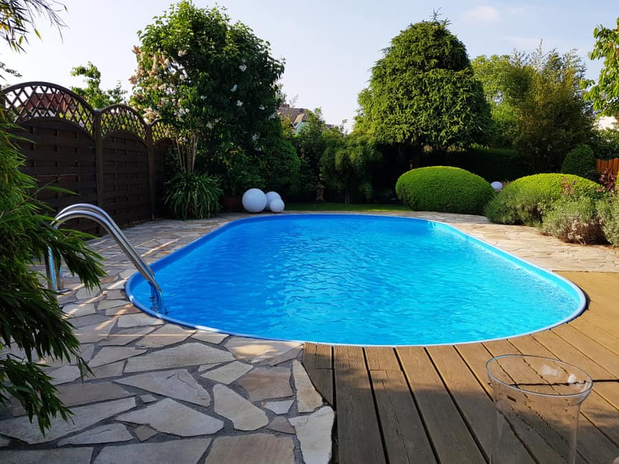 Backyard with a clear blue swimming pool, surrounded by stone and wooden decking, lush green bushes, and trees on a sunny day