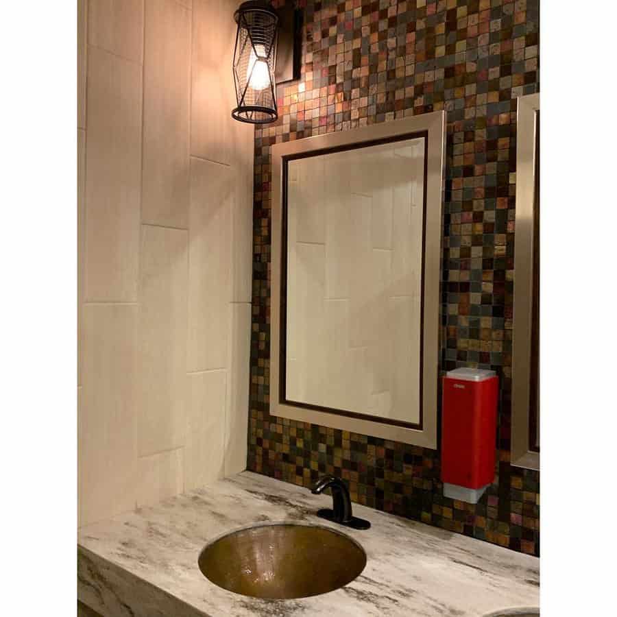 Industrial-style bathroom with multicolored stone tile backsplash, black faucet, round bronze sink, and exposed bulb wall sconce.