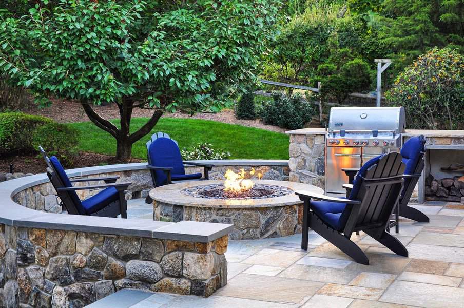 Outdoor patio with stone fire pit, surrounded by four black chairs with blue cushions, a grill stands nearby, and trees in the background