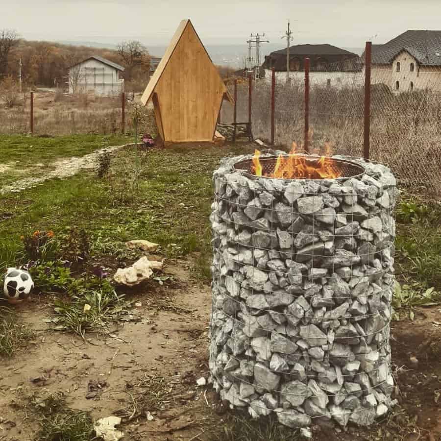 Stone fire pit with burning fire in a yard, a small wooden hut and a soccer ball nearby, overcast sky in the background