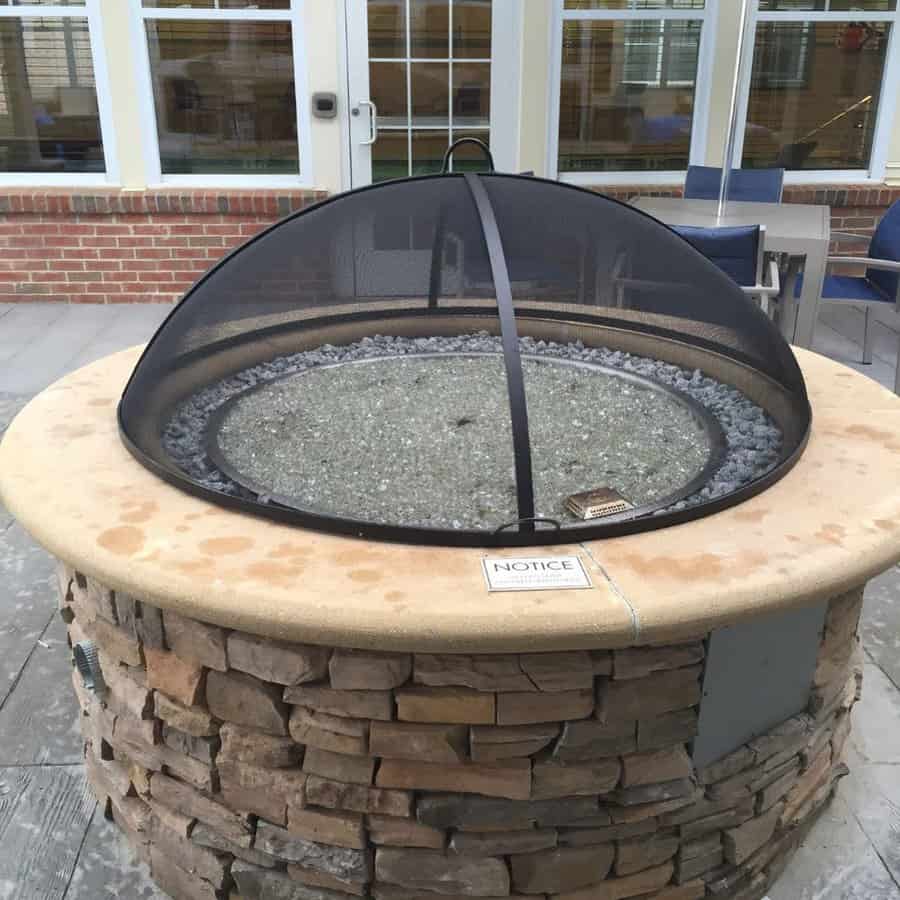 Stone fire pit with a mesh cover, surrounded by patio chairs, in front of windows, notice sign visible on its side