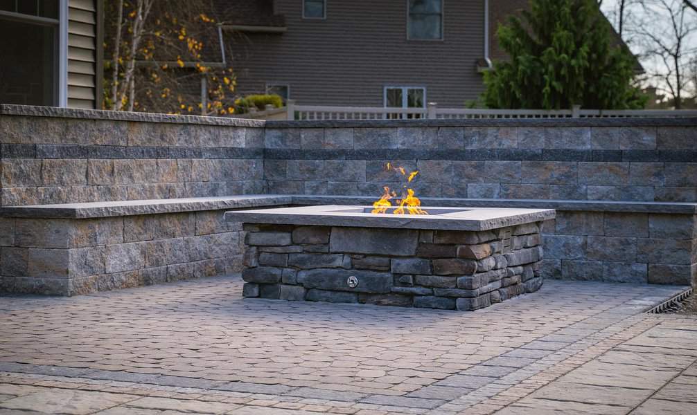 Outdoor stone fire pit with flames, surrounded by brick walls and pavement, in a backyard setting