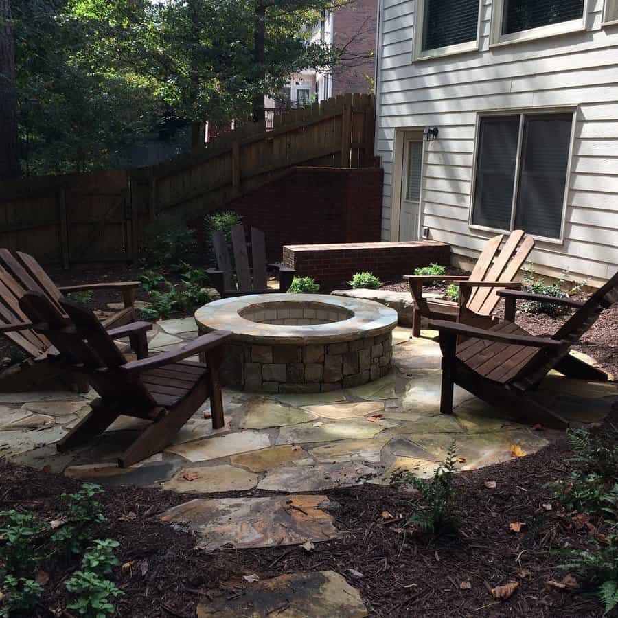 Outdoor patio with a stone fire pit and four wooden chairs, surrounded by a wooden fence and plants