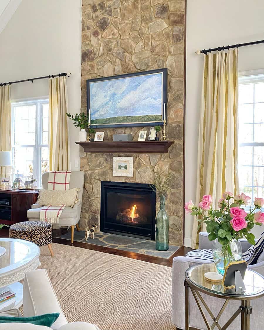 A rustic stone fireplace with a wooden mantel, framed by large windows, a colorful painting above, and elegant decor, creating a warm and inviting living space