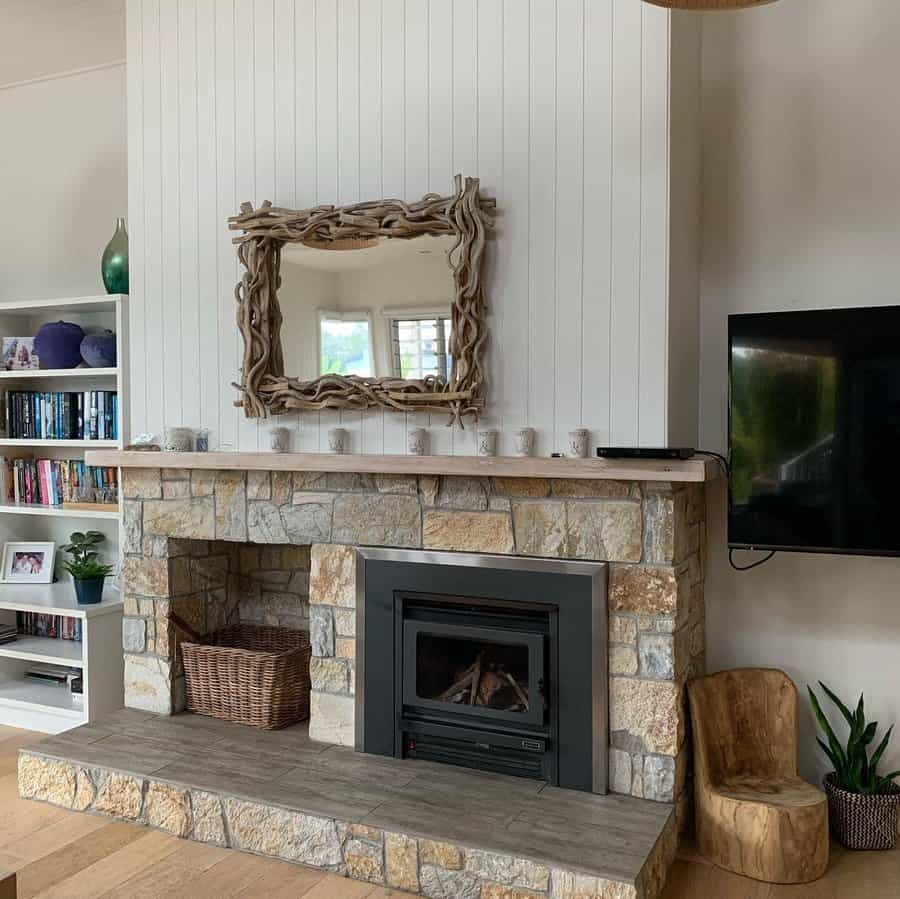 A rustic stone fireplace with a wooden mantel, framed by a driftwood mirror, a modern stove, and simple decor, offering a cozy, natural feel to the room