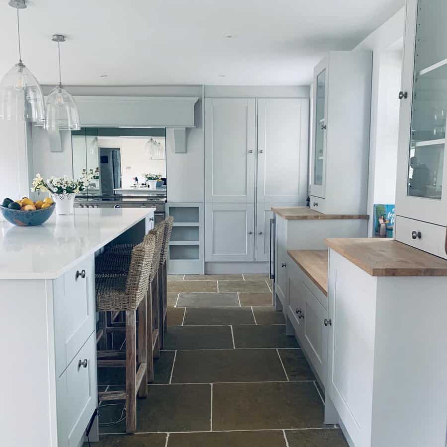 Modern kitchen with white cabinets, a large island with wicker stools, glass pendant lights, and stone tile flooring