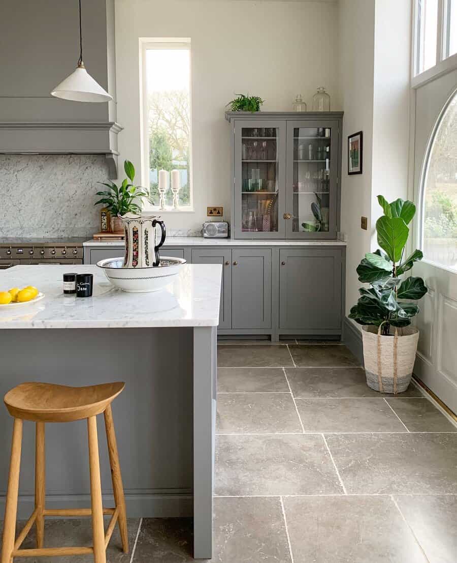 Large stone tile flooring enhances this elegant kitchen, complementing soft gray cabinetry, marble countertops, and warm wood accents for a timeless look