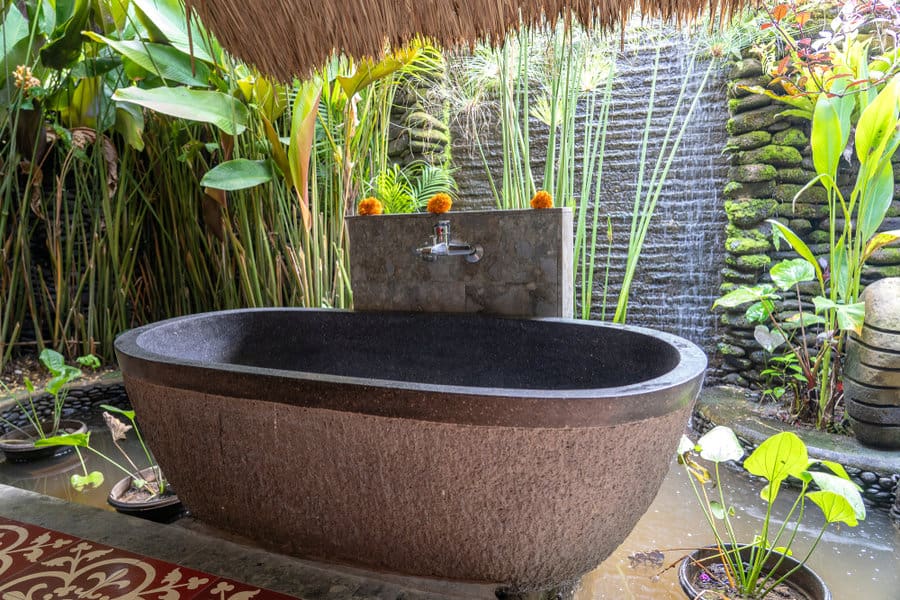 Stone bathtub in lush outdoor setting with green plants and a waterfall in the background