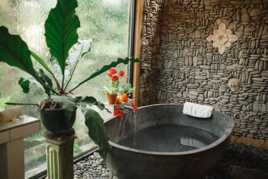 Stone bathtub in rustic bathroom with pebble wall, plants, and large window with garden view