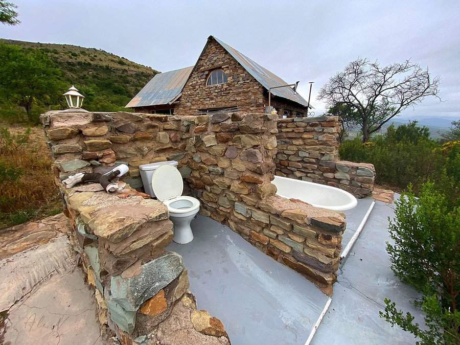 Outdoor bathroom with stone walls, featuring a toilet, sink, and bathtub; rustic stone house in the background, surrounded by nature