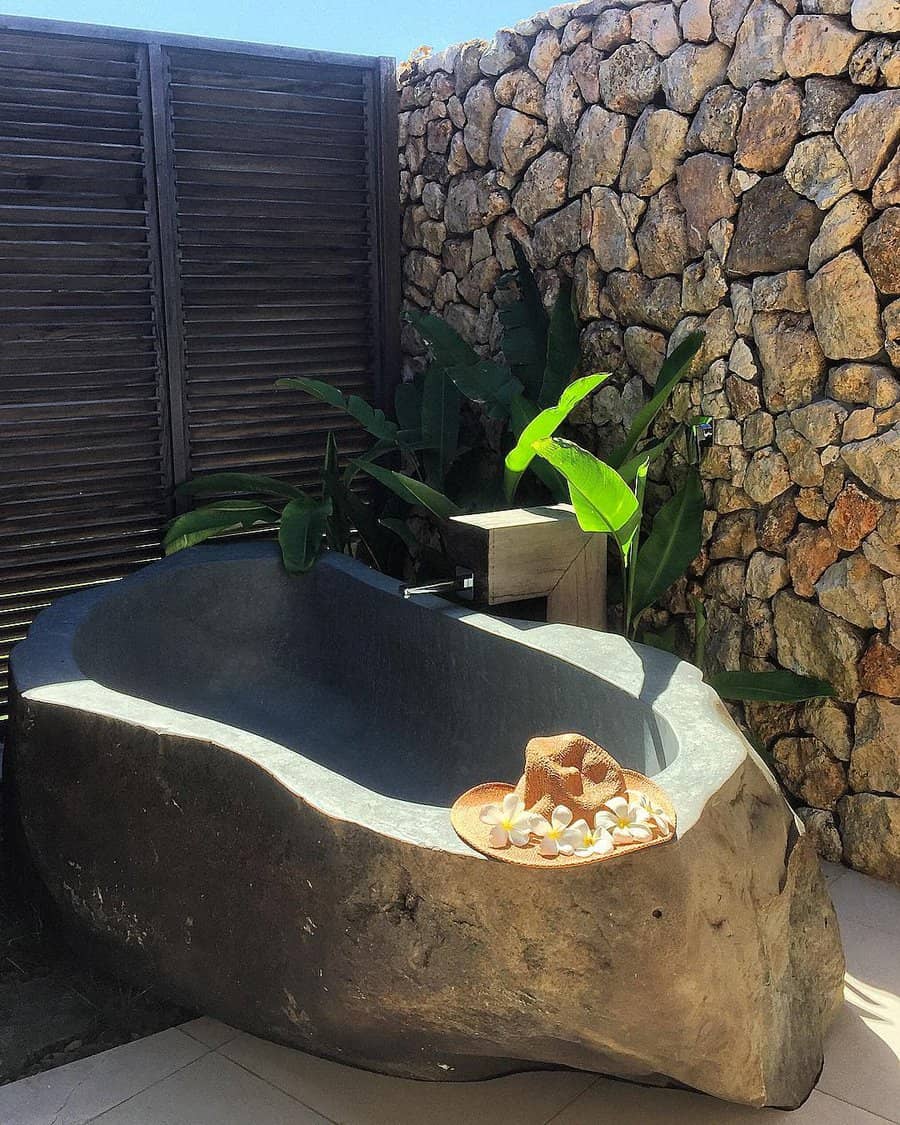 Stone bathtub outdoors with tropical plants, a rock wall, and wooden screen; sun hat with shells rests on the tub's edge