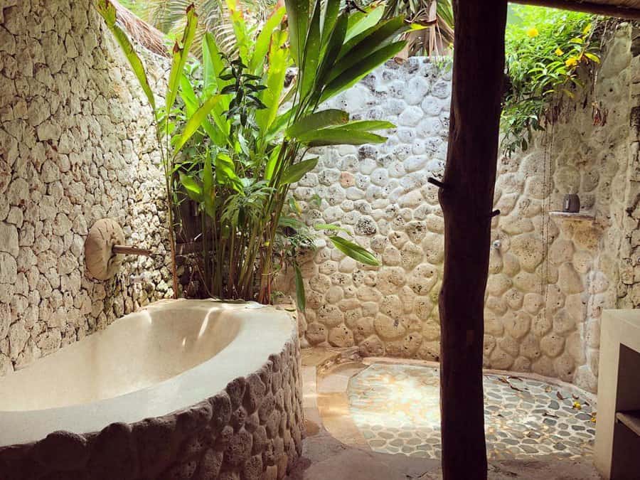 Outdoor stone bathroom with a bathtub and shower, surrounded by lush green plants and natural light filtering through