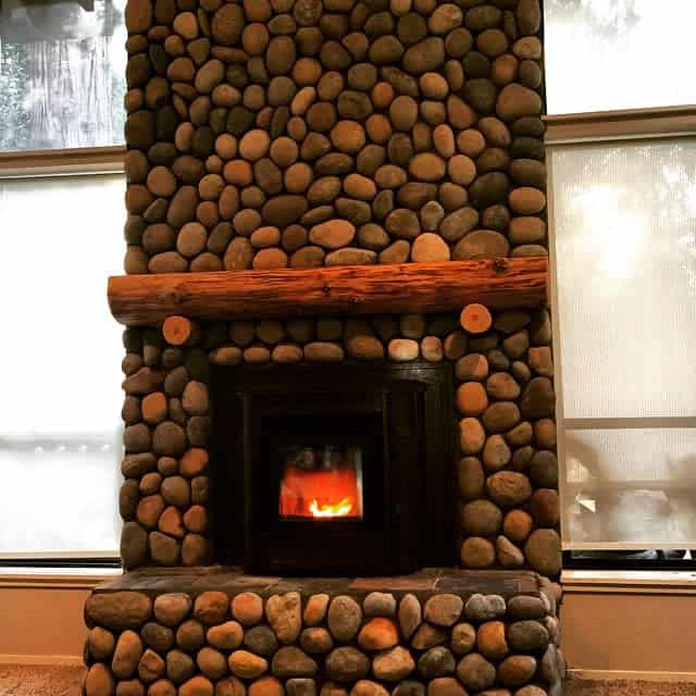 Rustic wood stove set within a floor-to-ceiling river rock hearth, featuring a thick wooden mantel for a cozy, nature-inspired ambiance.