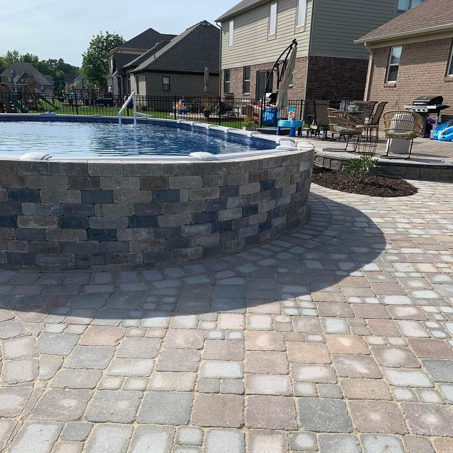 Above-ground pool with stone exterior on a paved patio, surrounded by chairs and houses under a clear sky