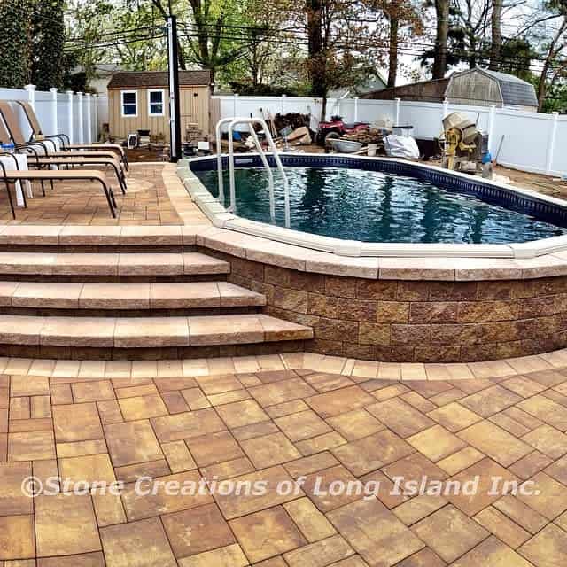 Elevated backyard pool with stone steps and patio, surrounded by a white fence and trees, with chaise lounges nearby