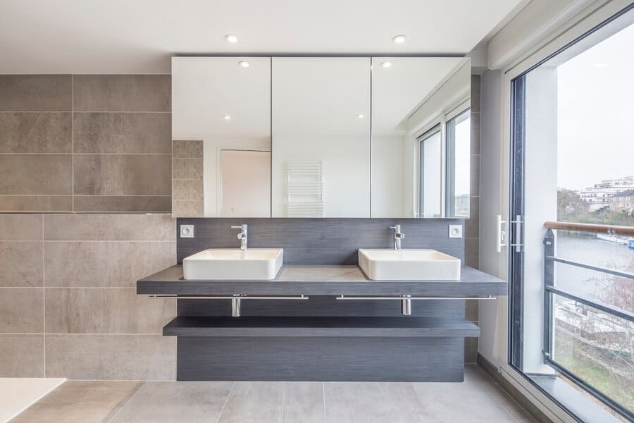 Modern bathroom with two sinks, large mirror, and window view of a river, featuring neutral tones and tiled walls for a sleek look