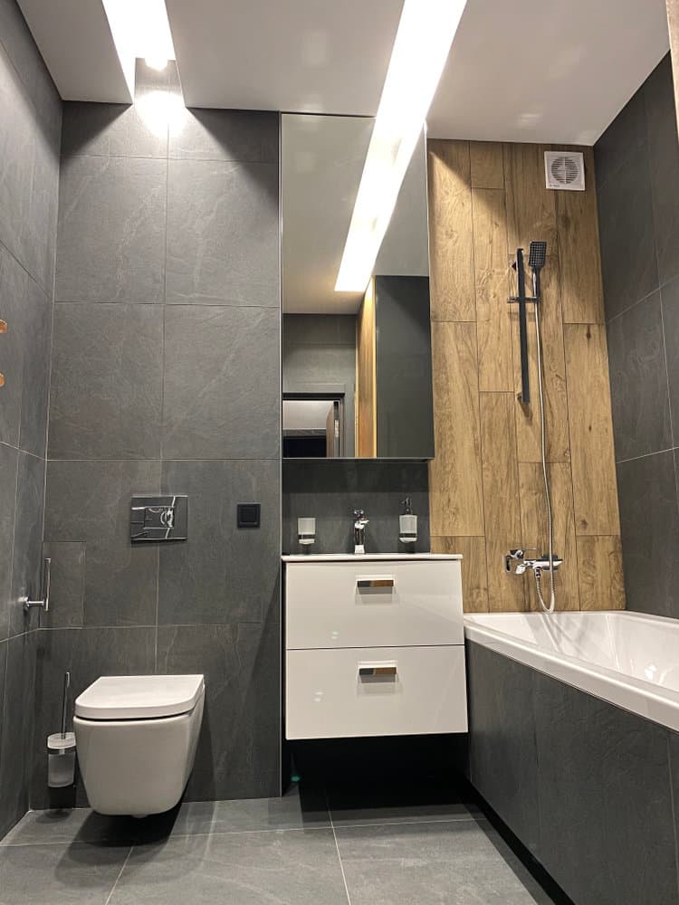 Modern bathroom with gray tiles, white wall-mounted toilet, vanity, mirror, and a bathtub with wood accent wall and shower head