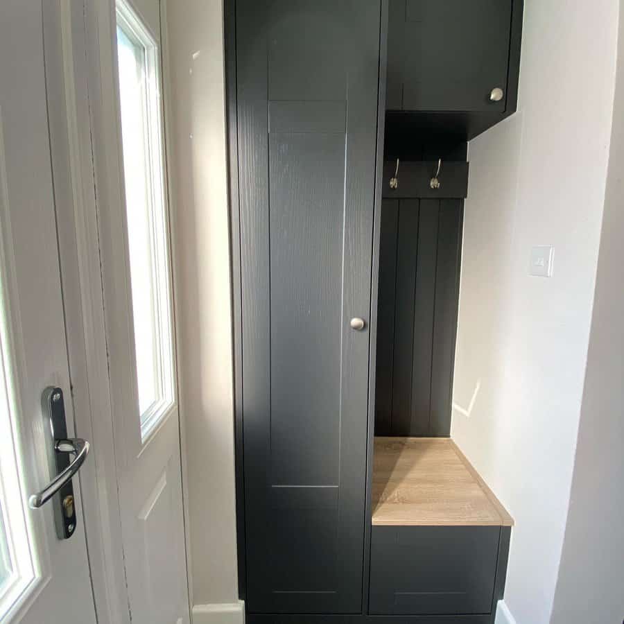 Dark gray hallway cabinet with a bench and hooks, next to a door with frosted glass, in a bright, narrow space