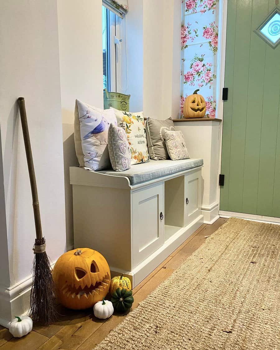 Cozy cottage entryway with a storage bench, floral accents, festive pumpkins, and a rustic broom, creating a charming autumn-inspired space