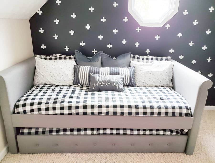 A cozy daybed with checkered bedding and pillows, against a black wall with white plus patterns, under a small hexagonal window