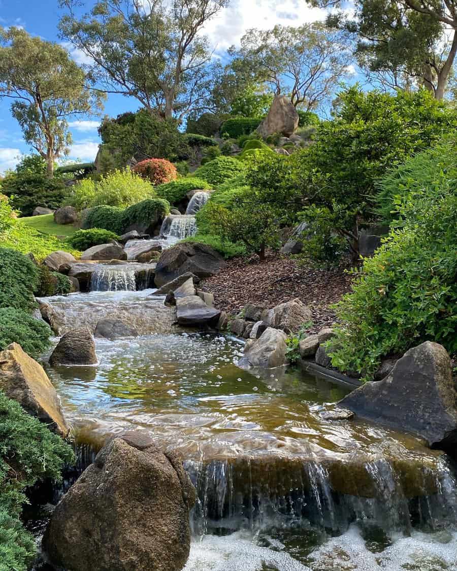 A serene waterfall flowing through a lush, green garden with rocks and trees under a partly cloudy sky