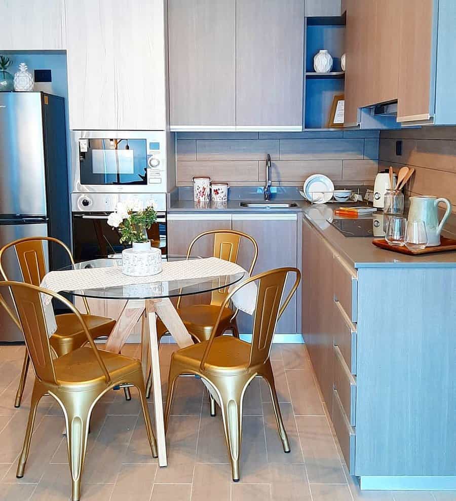 Modern kitchen with wooden cabinets, a round table with four gold chairs, stainless steel fridge, and appliances; decor includes a potted plant