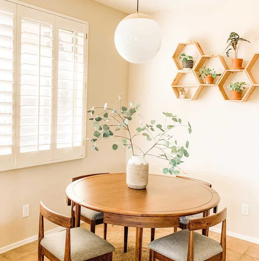 Bright dining area with a round wooden table, chairs, a vase with greenery, hexagonal shelves with plants, and a white pendant light