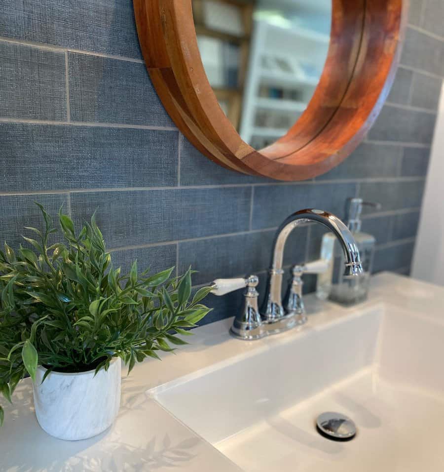 Modern bathroom with dark textured subway tile backsplash, chrome faucet, round wood mirror, and a marble planter with greenery.