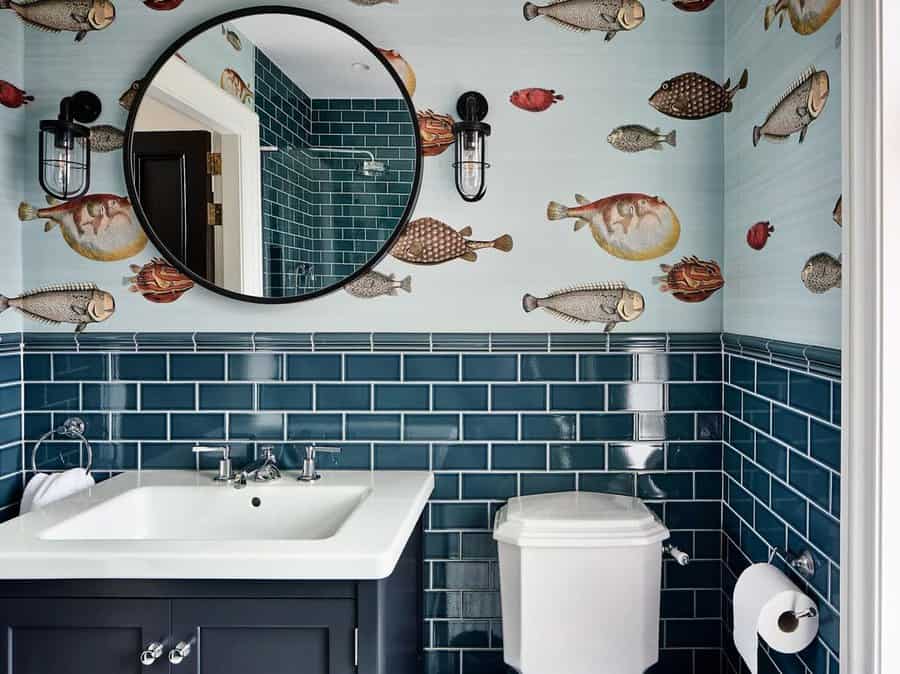 Playful bathroom with glossy dark teal subway tile backsplash, fish-themed wallpaper, round mirror, and industrial-style sconces.