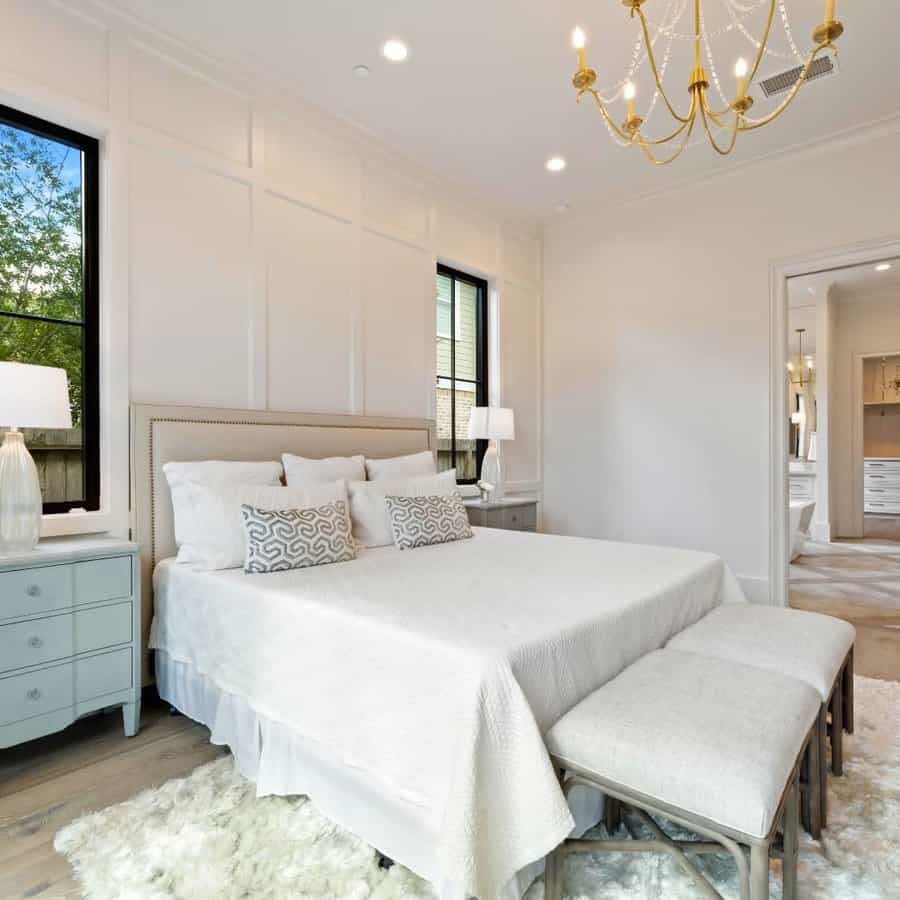 A bright bedroom with a king-size bed, plush rug, two benches, bedside tables, and a chandelier, with windows showing lush greenery