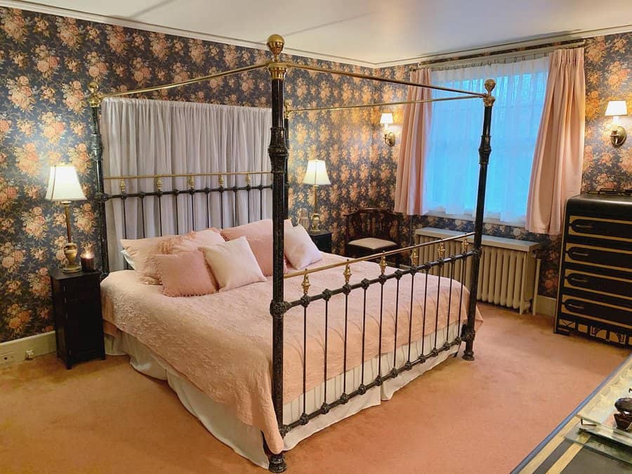 Master bedroom with a vintage four-poster bed, floral wallpaper, pink bedding, curtains, warm lighting, and a dresser