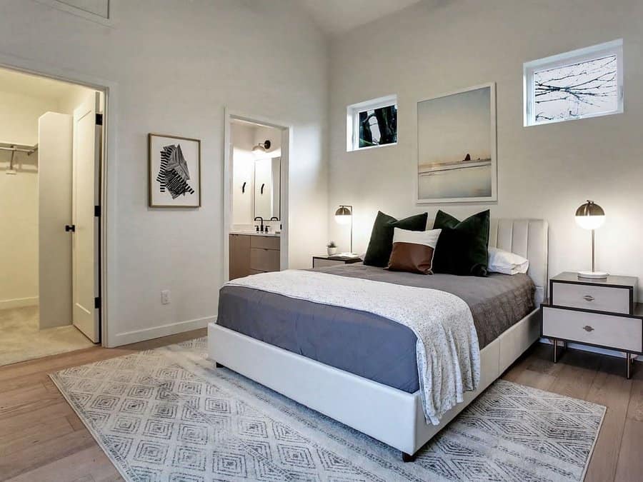 Modern bedroom with gray bedding, white bedframe, two windows, artwork, bedside tables with lamps, and view into a closet and bathroom