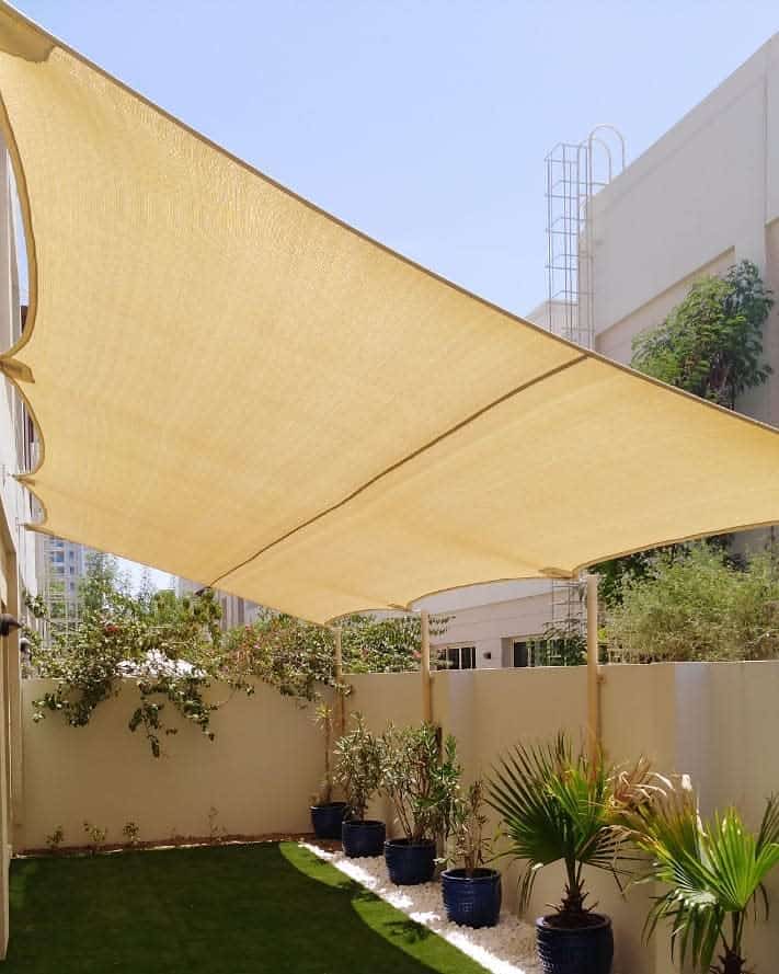 Small backyard with a beige shade sail, artificial grass, potted plants, and a white pebble border for a shaded outdoor retreat