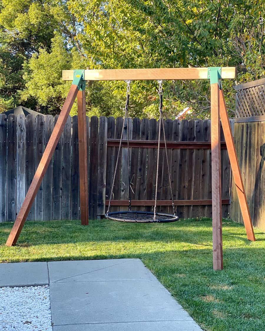 A wooden swing set with a round net swing hangs in a grassy backyard, enclosed by a wooden fence and leafy trees