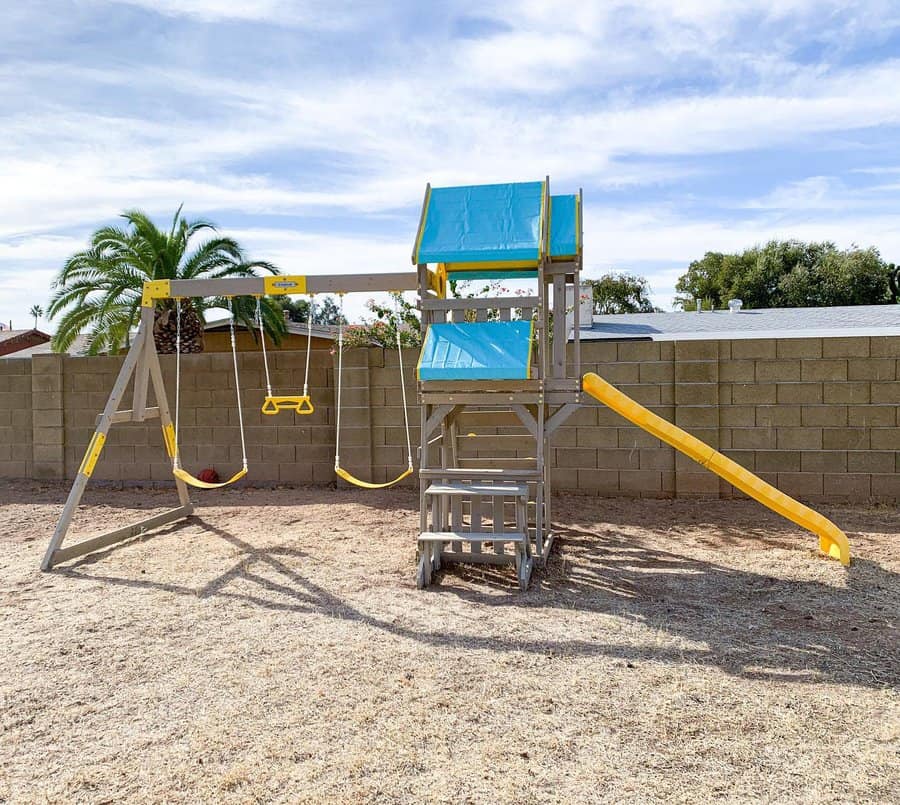 Wooden playground set with swings, slide, and climbing platform, palm tree and brick wall in the background under a blue sky