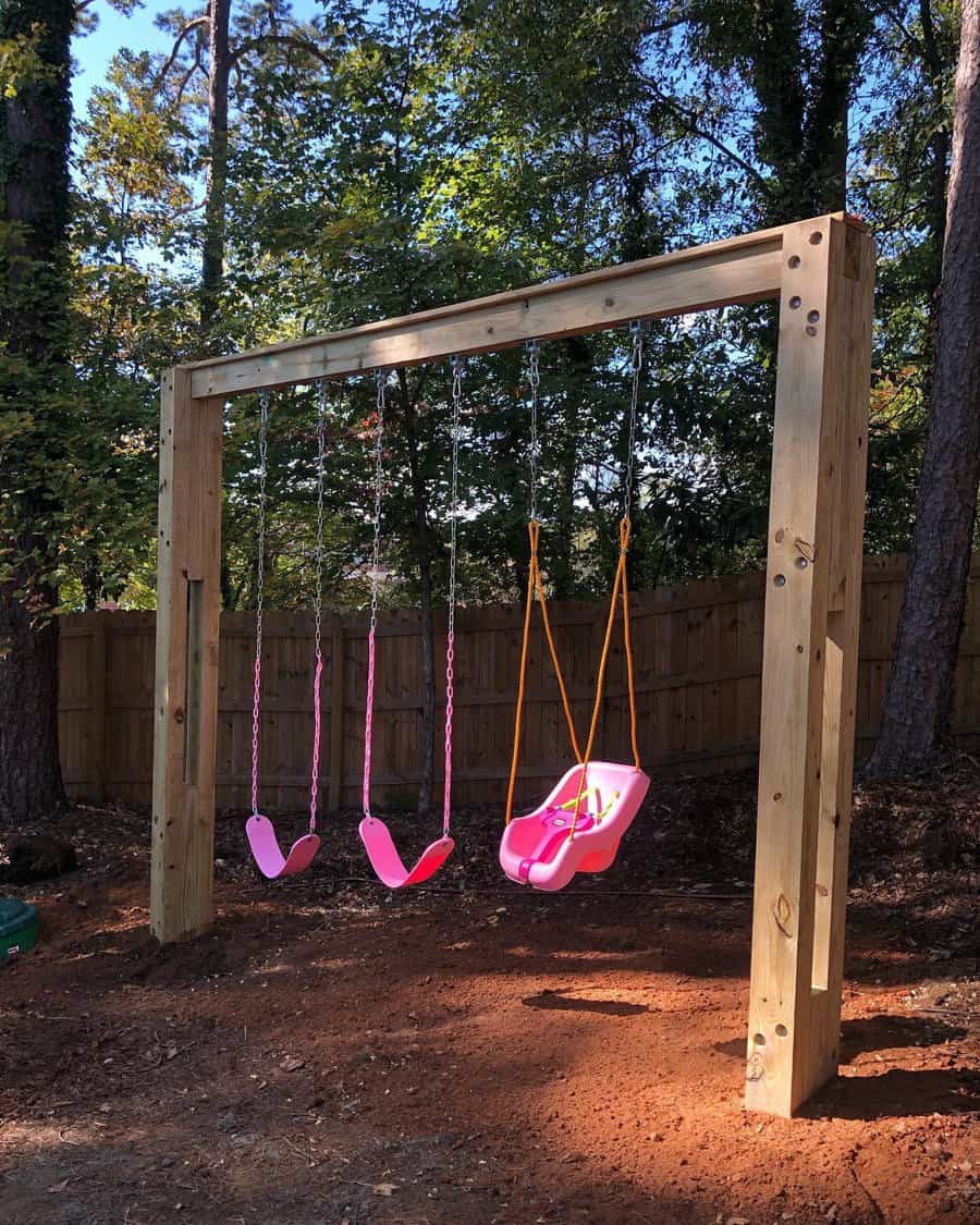 A wooden swing set with two pink swings and one pink baby swing in a backyard surrounded by trees and a wooden fence