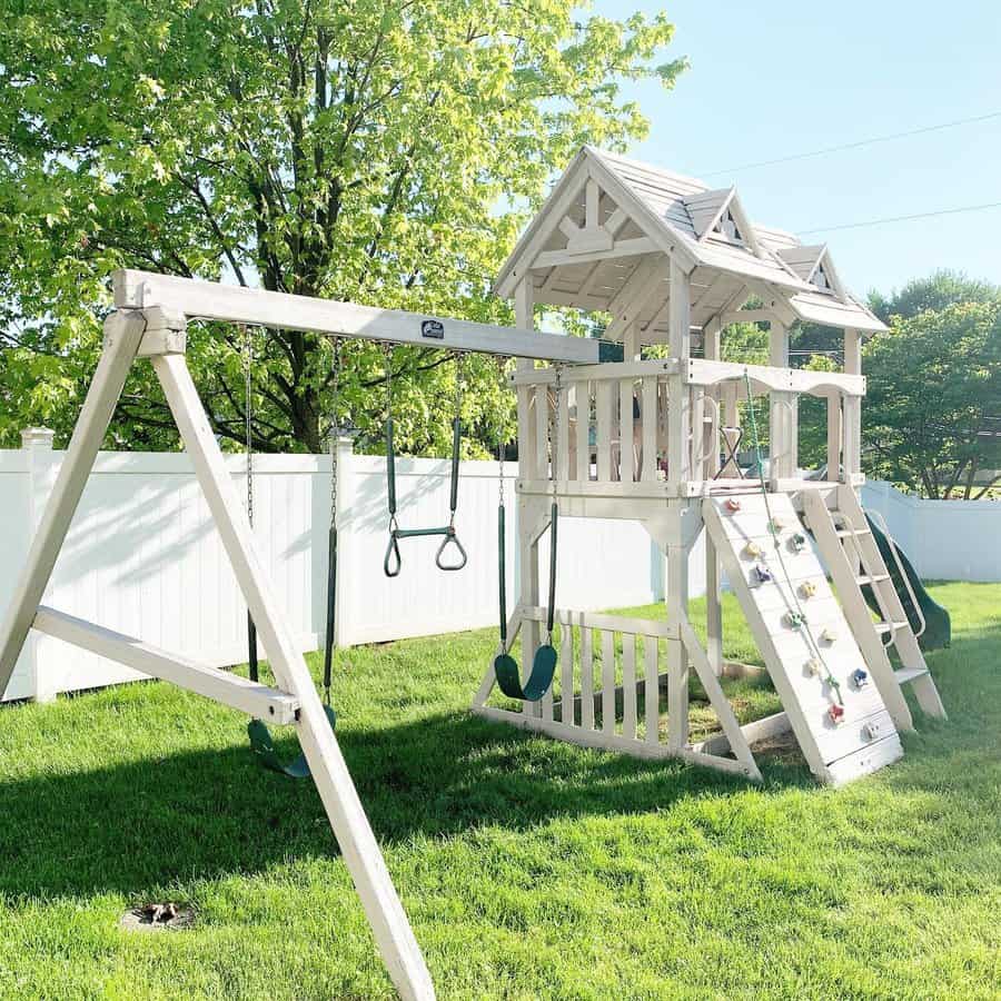 White wooden playset with swings, slide, and climbing wall on green grass, set against a sunny backdrop with a tree and white fence