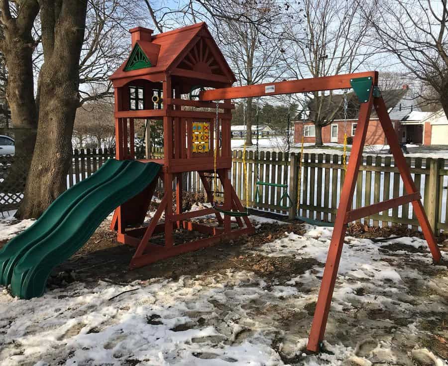 Wooden backyard playground with a green double slide, swing set, and tic-tac-toe panel, set in a snowy yard with a picket fence