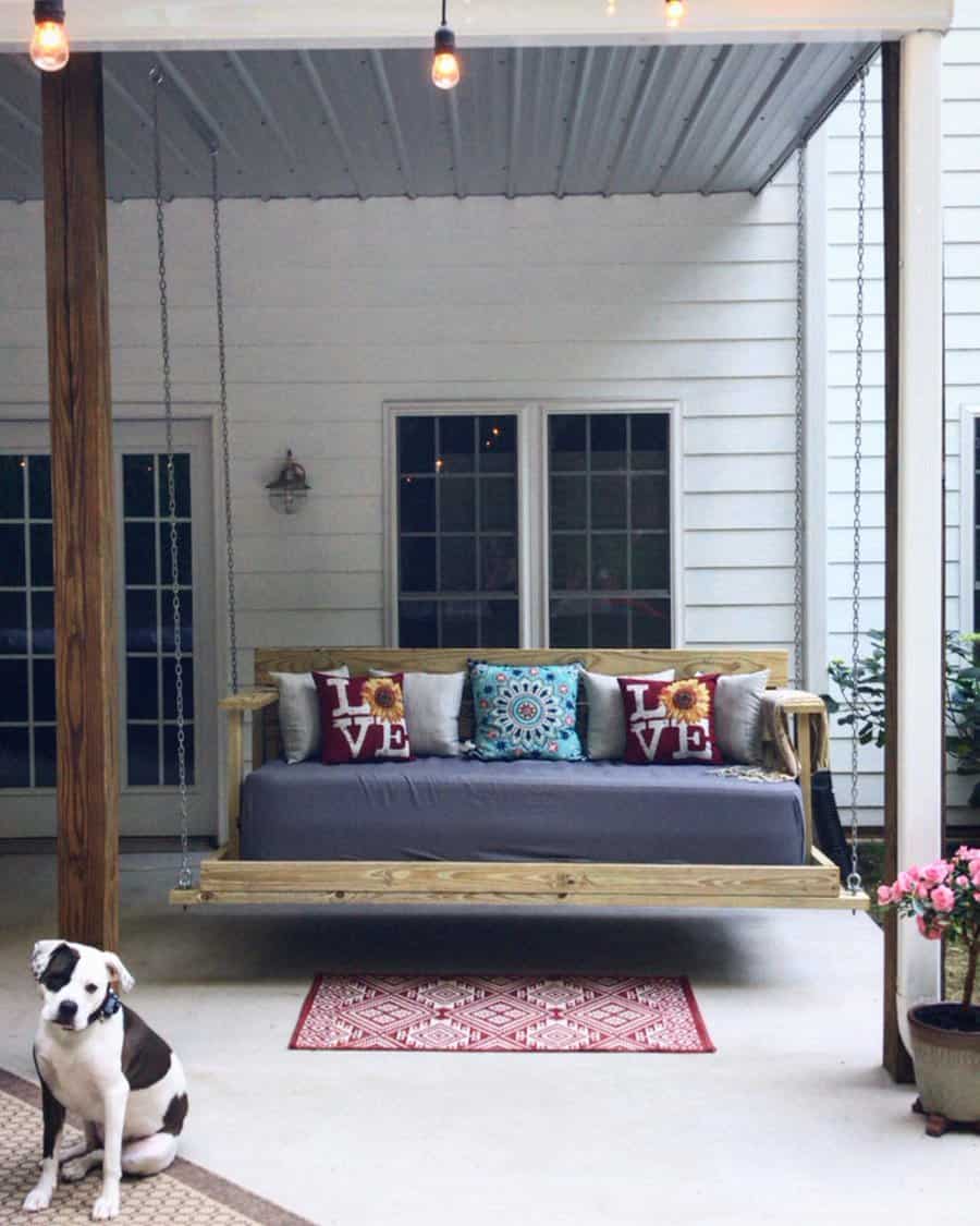 Swing daybed with colorful pillows on a patio, beneath string lights; a dog sits beside a potted plant on a red rug in the foreground