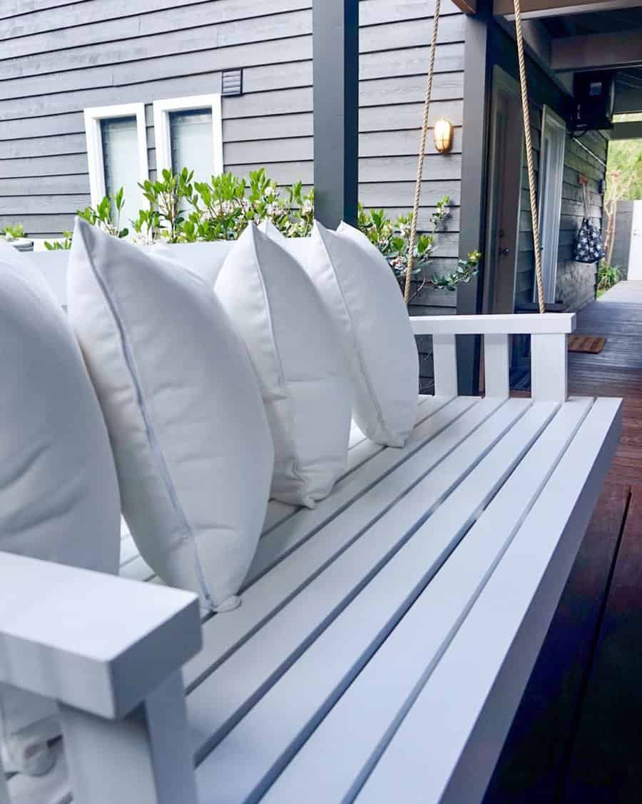 White outdoor swing bench with four white pillows on a wooden deck, surrounded by greenery and a gray house