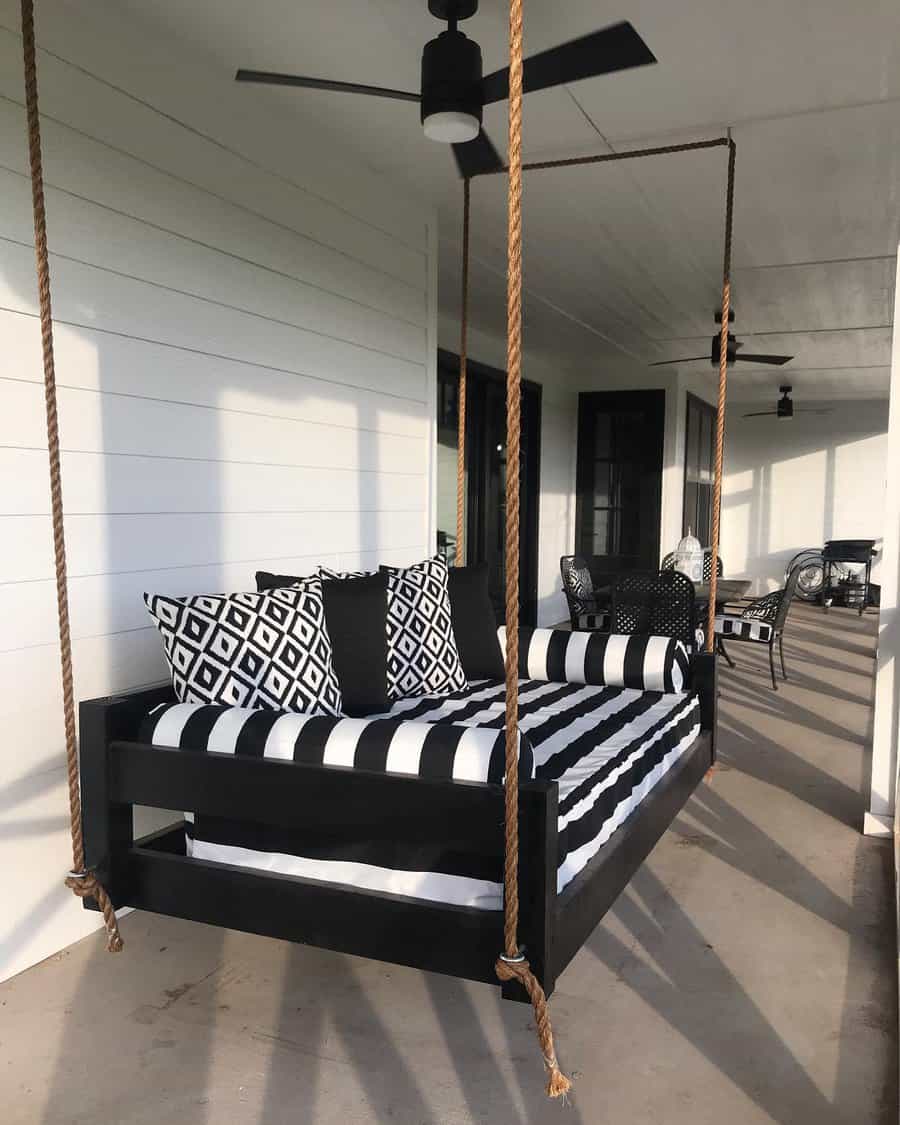 Swinging daybed with black and white striped cushions and geometric pillows on a porch, suspended by thick ropes, ceiling fan above