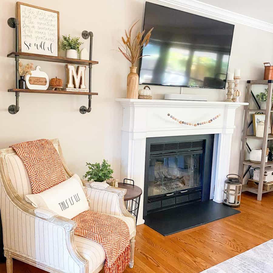 A modern white fireplace with a TV mounted above, decorated with a rustic garland and cozy accents, complemented by a comfortable armchair and shelving with plants and decor