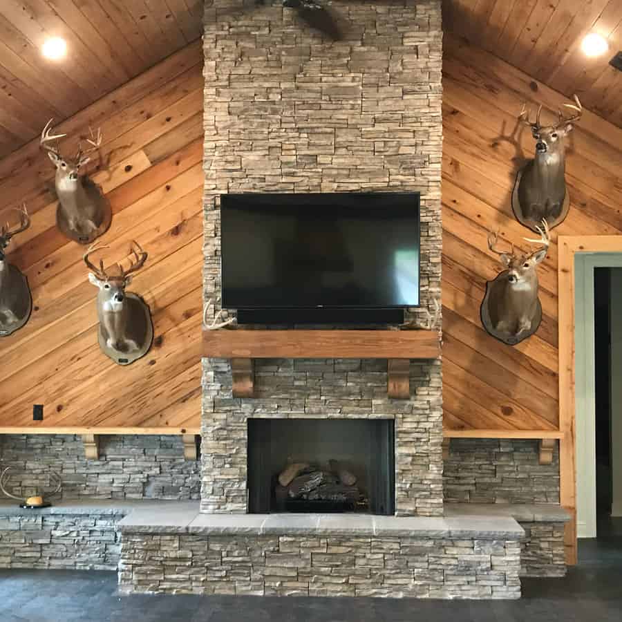 A rustic stone fireplace with a wooden mantel and mounted TV above, flanked by decorative taxidermy and wooden paneling for a cozy, cabin-like feel