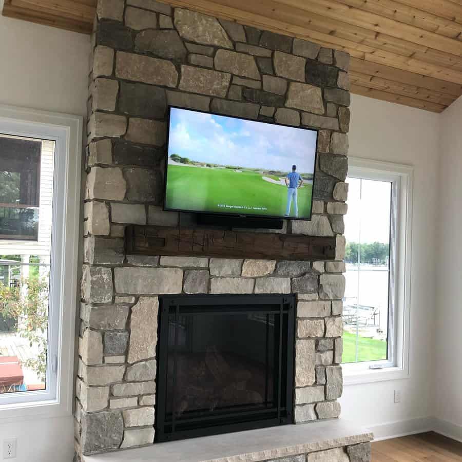 A rustic stone fireplace with a wooden mantel, featuring a wall-mounted TV above and a sleek black frame, offering a cozy yet modern look