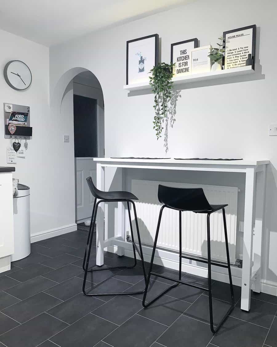 Minimalist kitchen nook featuring a tall white table, two modern black stools, wall decor, hanging plant, and a dark tiled floor