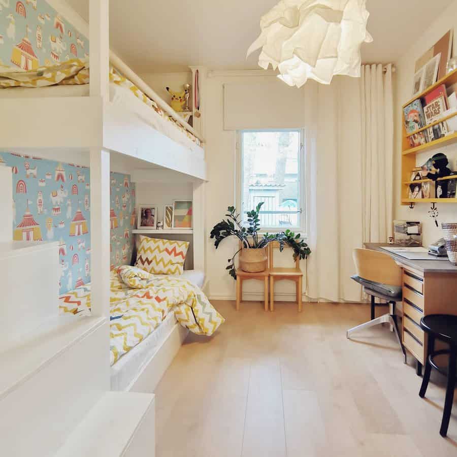 Children's room with bunk beds, colorful wallpaper, a desk with shelves, two small chairs, and a window with a leafy view