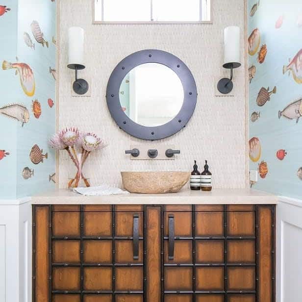 Coastal-themed bathroom with textured cream backsplash, vessel sink, round porthole-style mirror, and playful fish wallpaper on side walls.