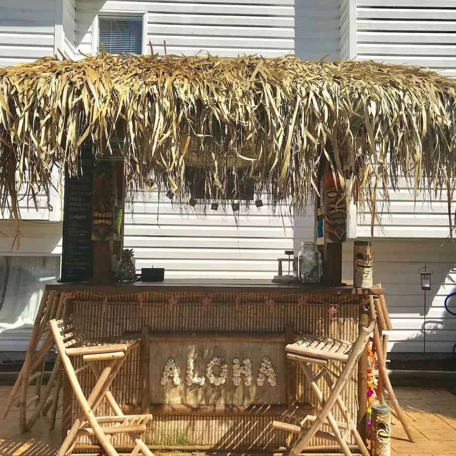 Tiki bar with thatched roof, bamboo stools, and "Aloha" sign, set against a white building background