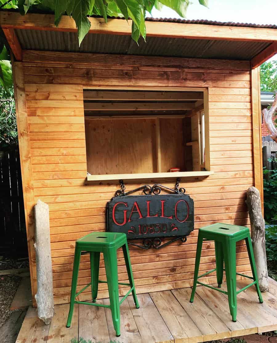 A small wooden outdoor bar with a sign "Gallo 1055" and two green stools in front